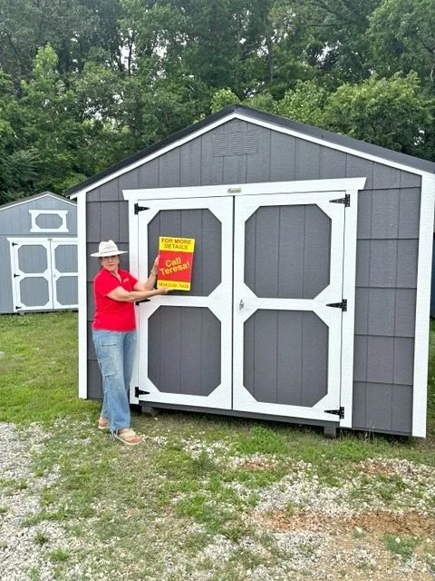 Woman in red shirt and hat holding a sign in front of a gray shed with white garage doors.