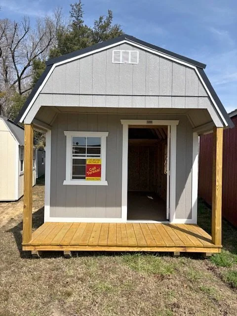 A small gray shed with a gable roof, an open front door, a window with a 'For Sale' sign, and a wooden porch in front.