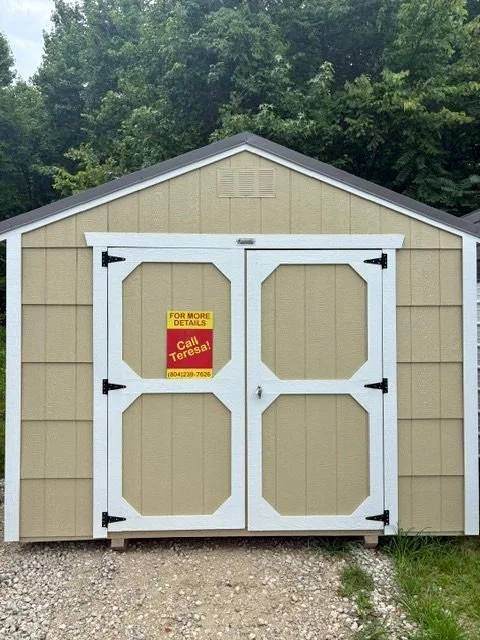 A small beige storage shed with white double doors and black hinges, situated outdoors on a gravel surface with trees in the background. There is a yellow and red 'For More Details' sign on the left door with a phone number.