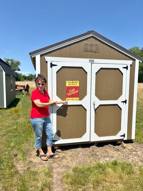 Woman standing next to a small shed with a sign that says "For More Details Call Teresa" and a phone number, outdoors on a sunny day.