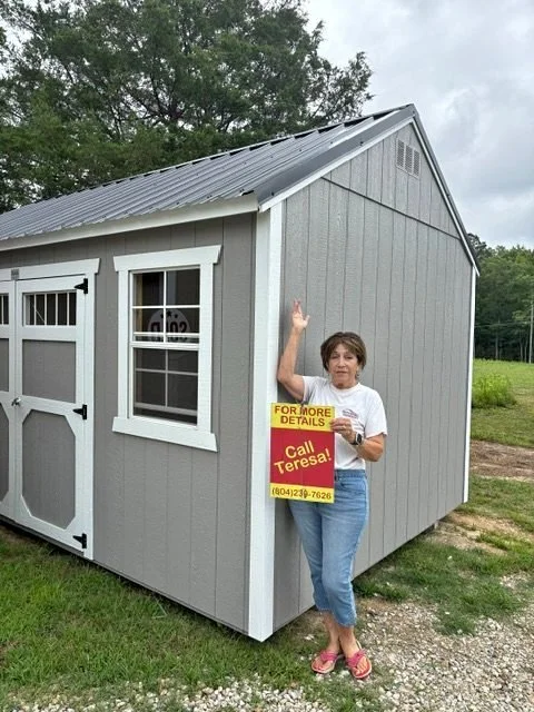 A woman standing next to a small gray shed holding a sign that reads 'For More Details Call Teresa!' with a phone number, waving with her right hand.