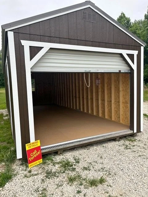 Open brown storage shed with unfinished interior and a closed white roll-up door, situated outdoors on gravel with a yellow and red sign in front.