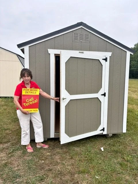 A woman standing outside a small shed holding a sign that says 'For More Details Call Teresa' with a phone number, pointing towards the open shed door.