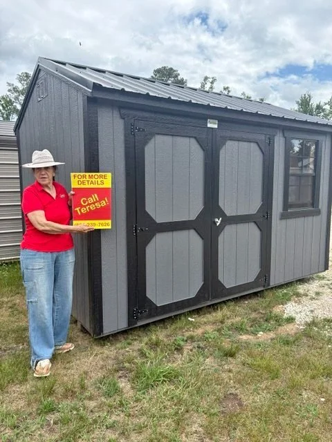 Woman in red polo shirt and wide-brim hat standing next to a gray and black shed holding a yellow sign that reads, 'For More Details Call Teresa!'