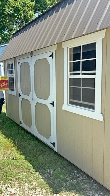 Exterior view of a beige shed with white trim and doors, a window with white trim, and a brown metal roof, situated on grass with a tree in the background.