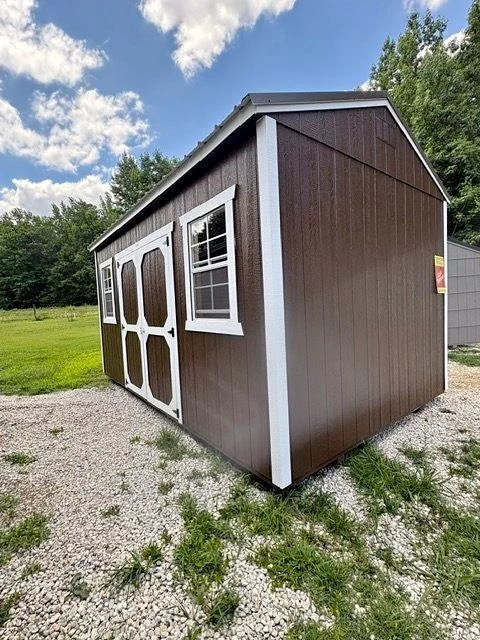 A brown shed with white trim, double doors, and two windows, situated on a gravel patch with grass and trees in the background.