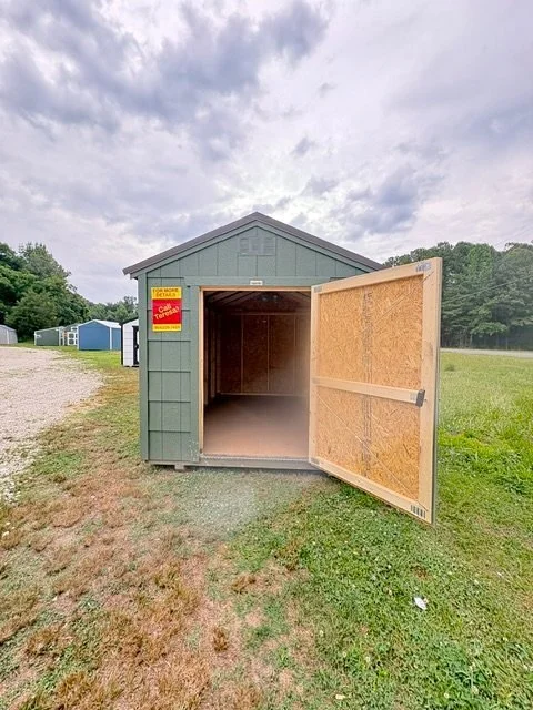 Small green storage shed with open door, situated on grassy area with overcast sky.