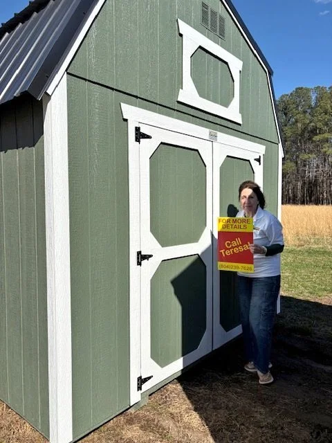 Woman standing outside green barn holding a yellow sign promoting a local real estate agent.