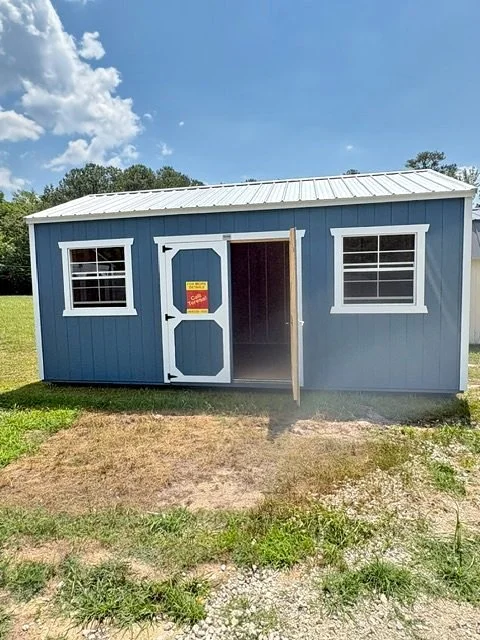 Blue shed with white trim, two windows, and an open door, on a grassy and gravel area under a partly cloudy sky.