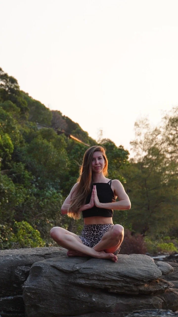 Eine Frau sitzt im Schneidersitz auf einem Felsen in einer Naturumgebung, beim Yoga im Sonnenuntergang.