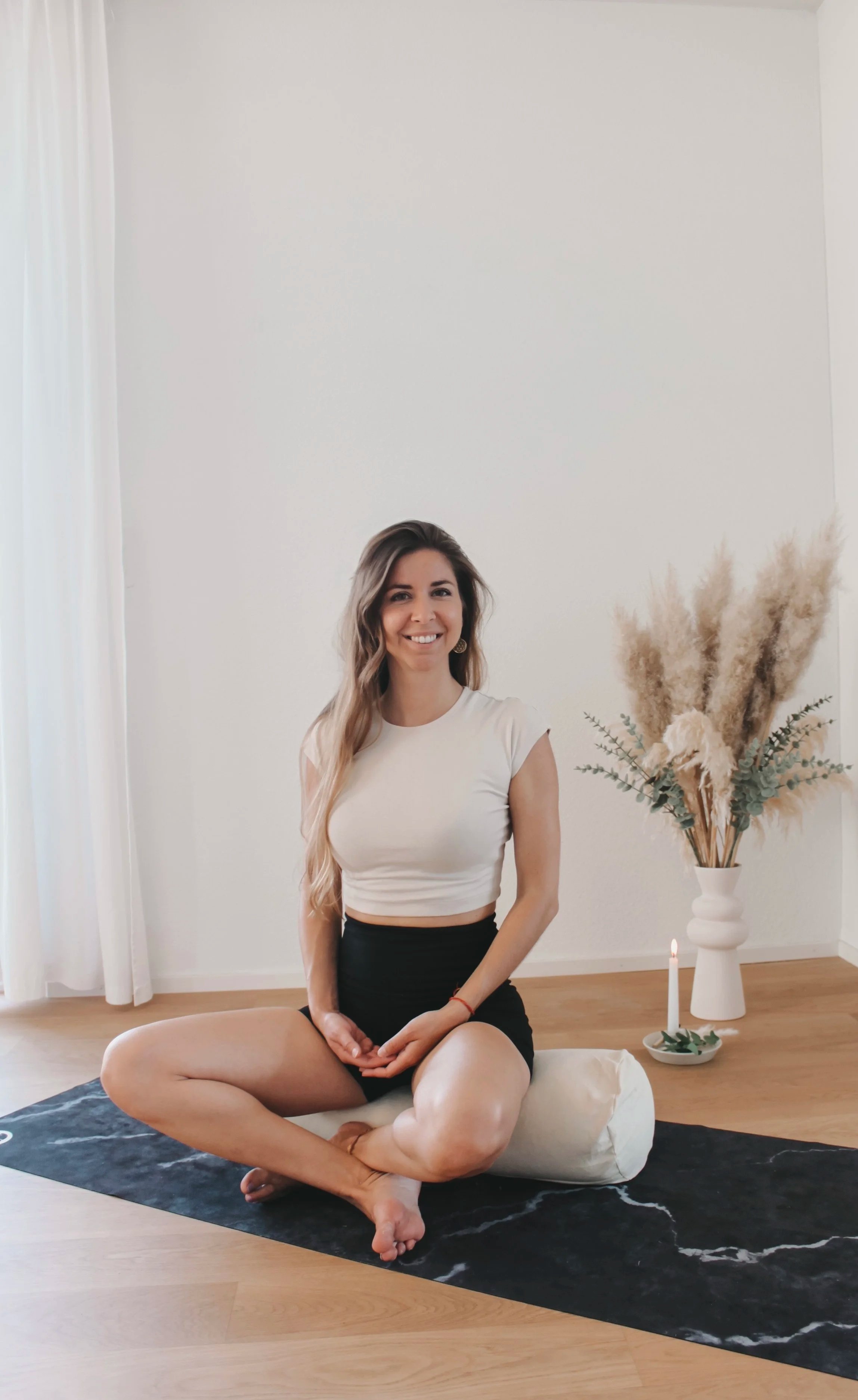Eine junge Frau sitzt in einer Meditationshaltung auf einer schwarzen Matte vor weißer Wand, neben einem weißen Kissen, mit einem weißen Kerzenleuchter und einem großen Blumenstrauß aus getrockneten Gräsern und Eukalyptus in einer weißen Vase im Hintergrund.