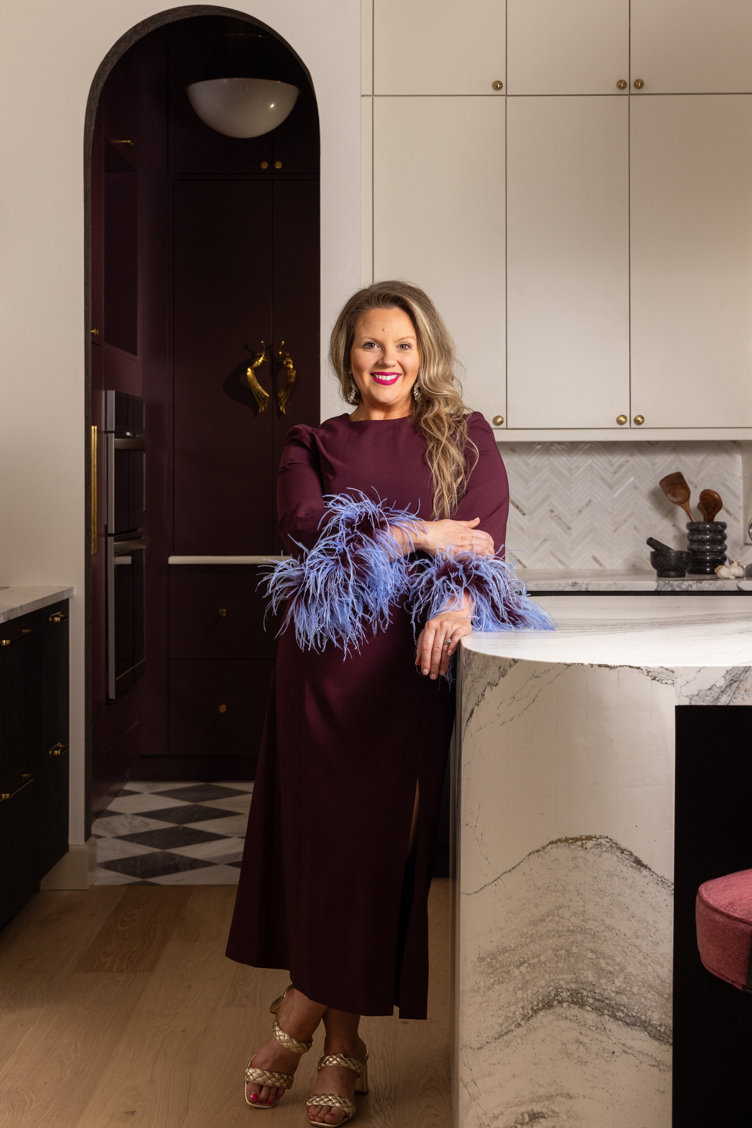 A woman in a maroon dress with feathered sleeves stands in a modern kitchen, smiling with arms crossed. She has long wavy hair and is wearing open-toe heels.