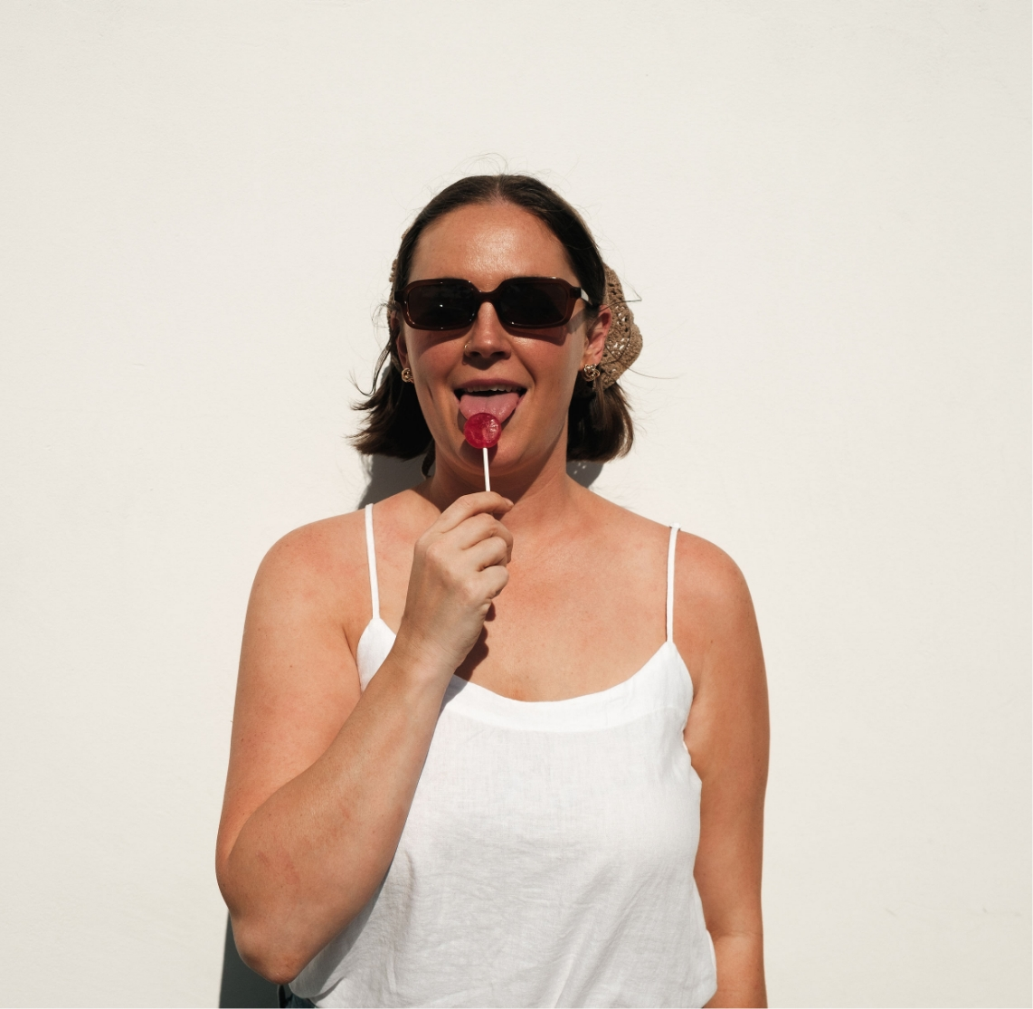 Woman wearing sunglasses and a white tank top holding a red lollipop against a plain white wall.