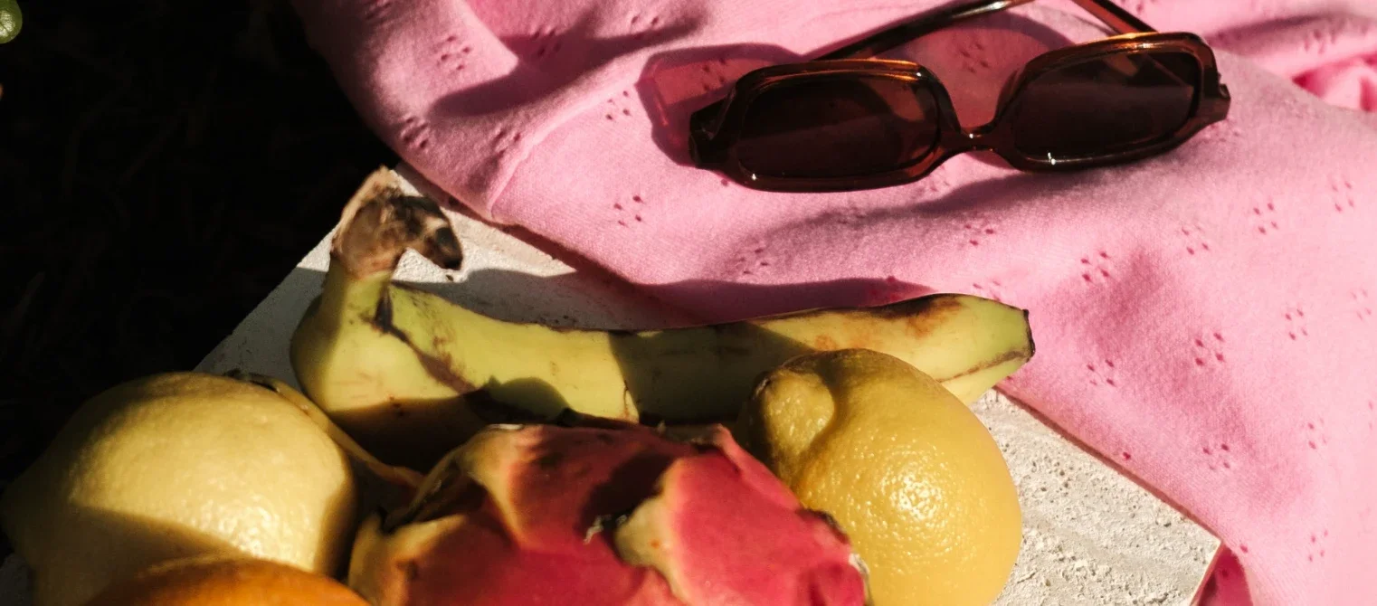 A pink cloth, a pair of sunglasses, and assorted fruits including lemons, a banana, and a dragon fruit, on a white surface.