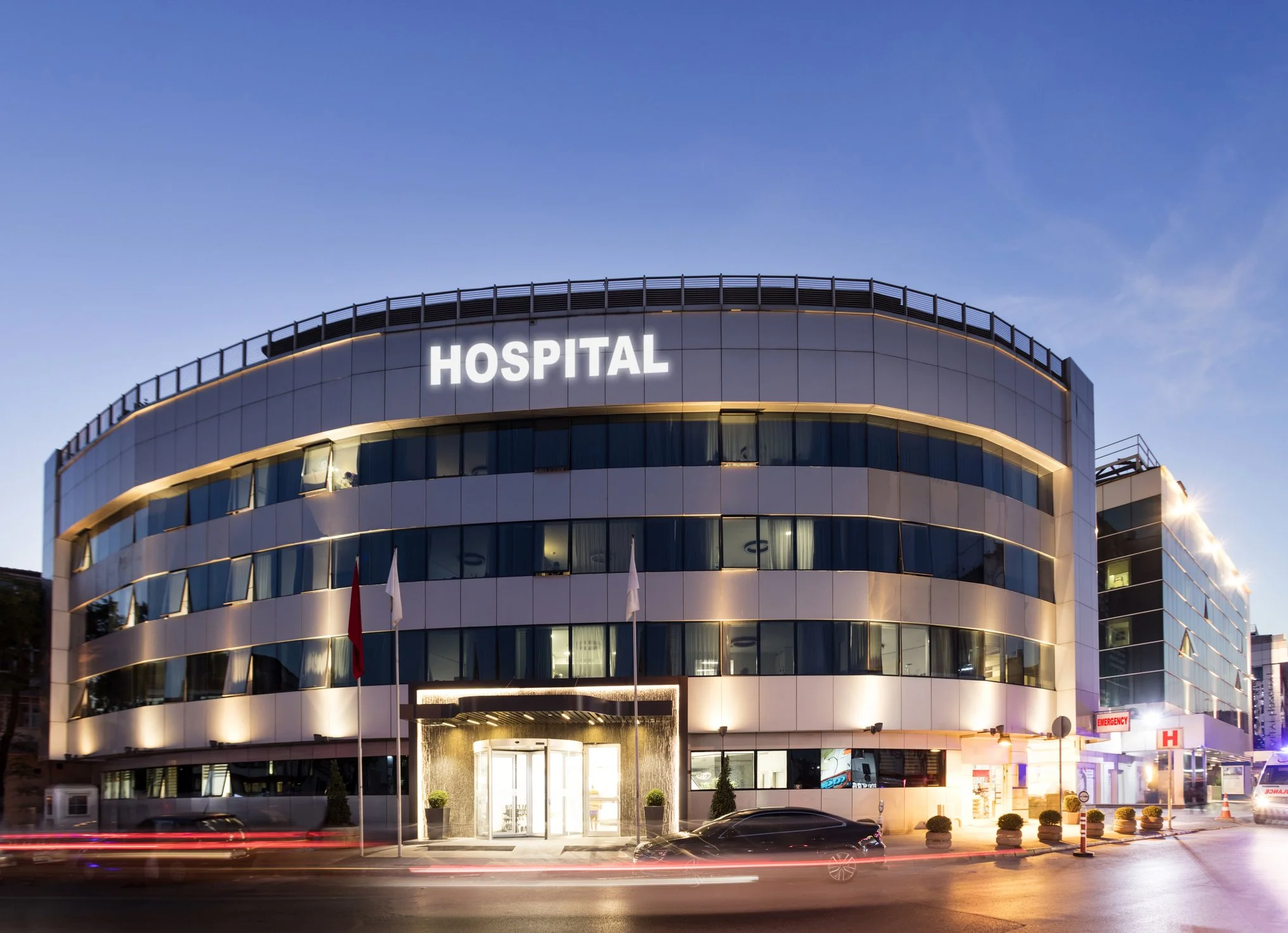 Modern multi-story hospital building at dusk with illuminated sign reading 'HOSPITAL' on the front and emergency entrance to the right.