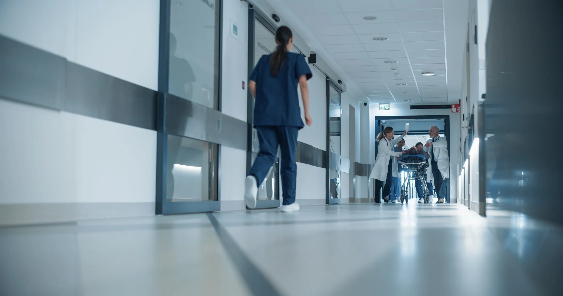 Hospital corridor with healthcare professionals attending to a patient on a stretcher, and a nurse walking down the hallway.