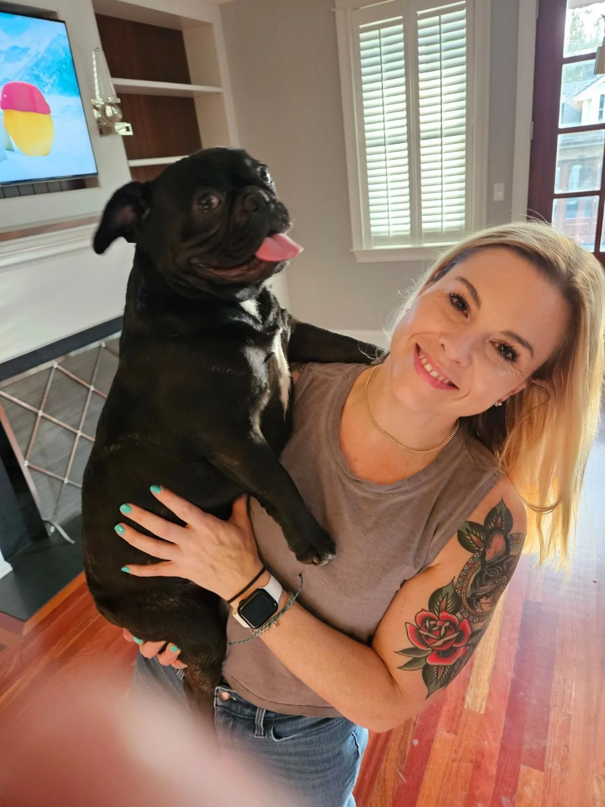 A woman standing in a living room holding a black pug. The woman has blonde hair, a sleeveless gray top, and a colorful tattoo on her arm depicting a rose around a dog's face. She is smiling at the camera, and there's a TV in the background showing a colorful image.
