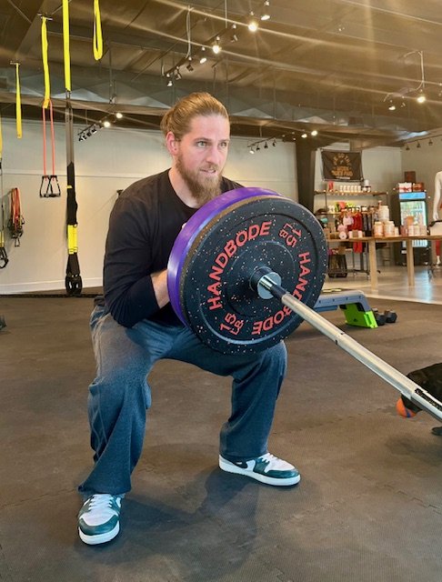 A man performing a squat with a barbell in a gym. He is holding the barbell placed in a landmine position, wearing casual athletic clothing and sneakers. The gym has various equipment in the background.