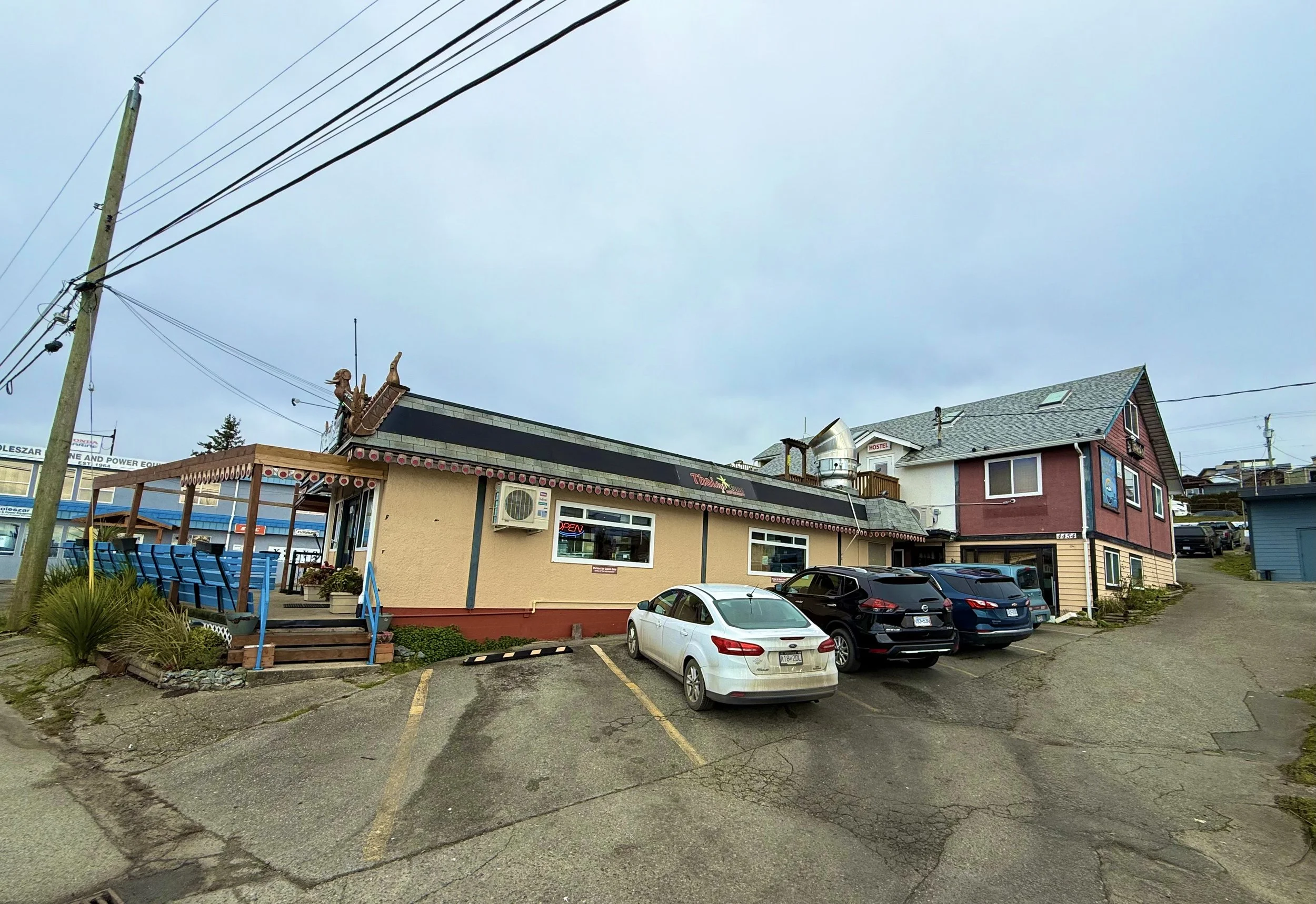 A small restaurant building with a porch and a parking lot, with cars parked in front, and a house-style building behind it, under an overcast sky.