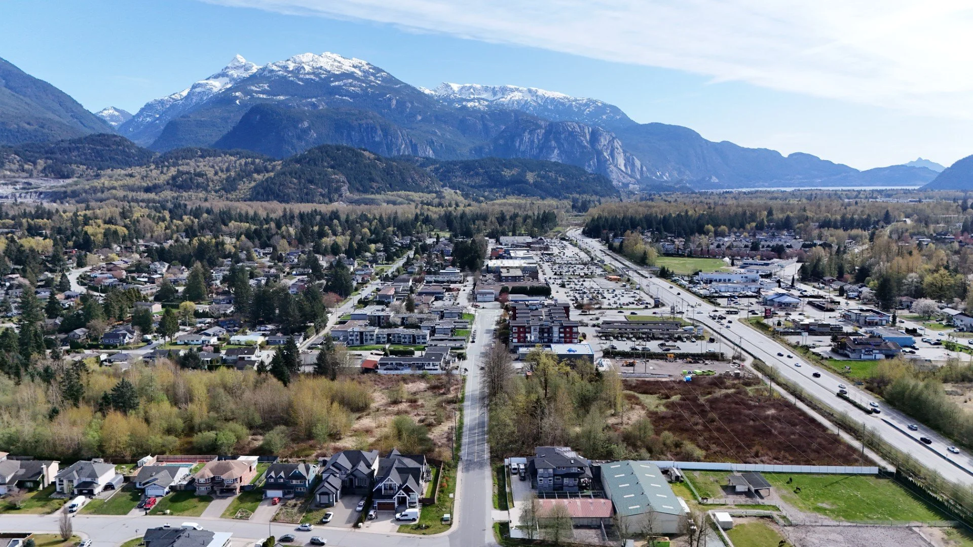 Aerial view of a small town at the base of snow-capped mountains, with residential houses, commercial buildings, and a main road with traffic.