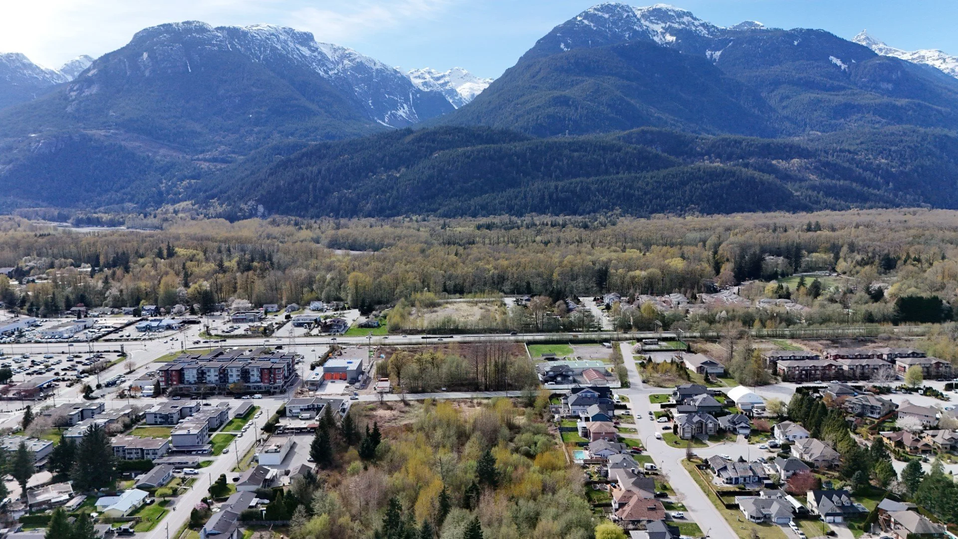 Aerial view of a suburban neighborhood with houses, roads, and a park, set against a backdrop of green mountains with snow-capped peaks.