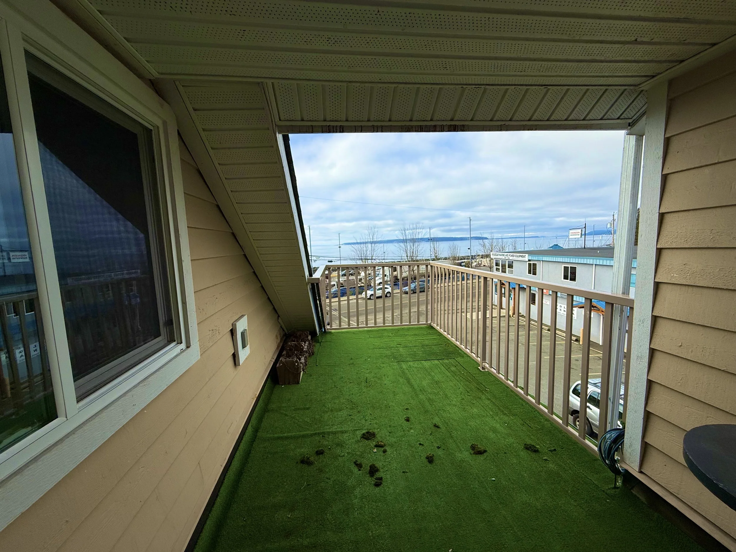 A small outdoor balcony with a green artificial grass floor, beige siding walls, and a white railing. There's a window on the left wall, a flower pot in the corner, and a view of parking lot and distant mountains under cloudy sky.