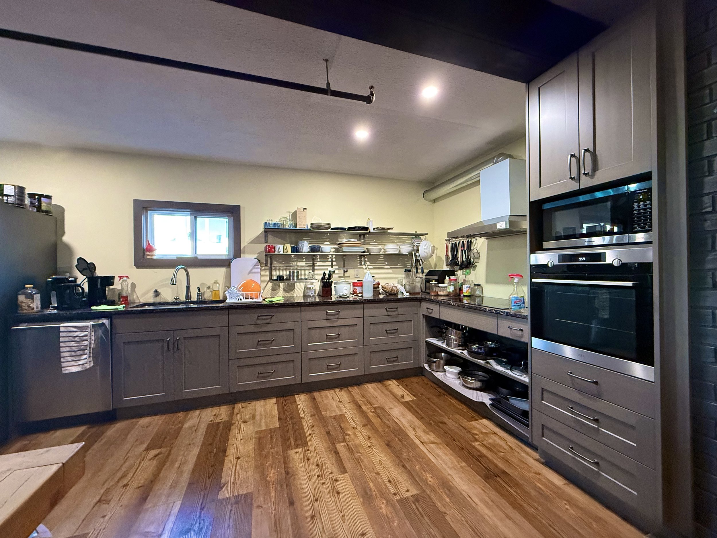 A kitchen with gray cabinets and wood flooring, featuring a small window, a black refrigerator, and a built-in oven and microwave.