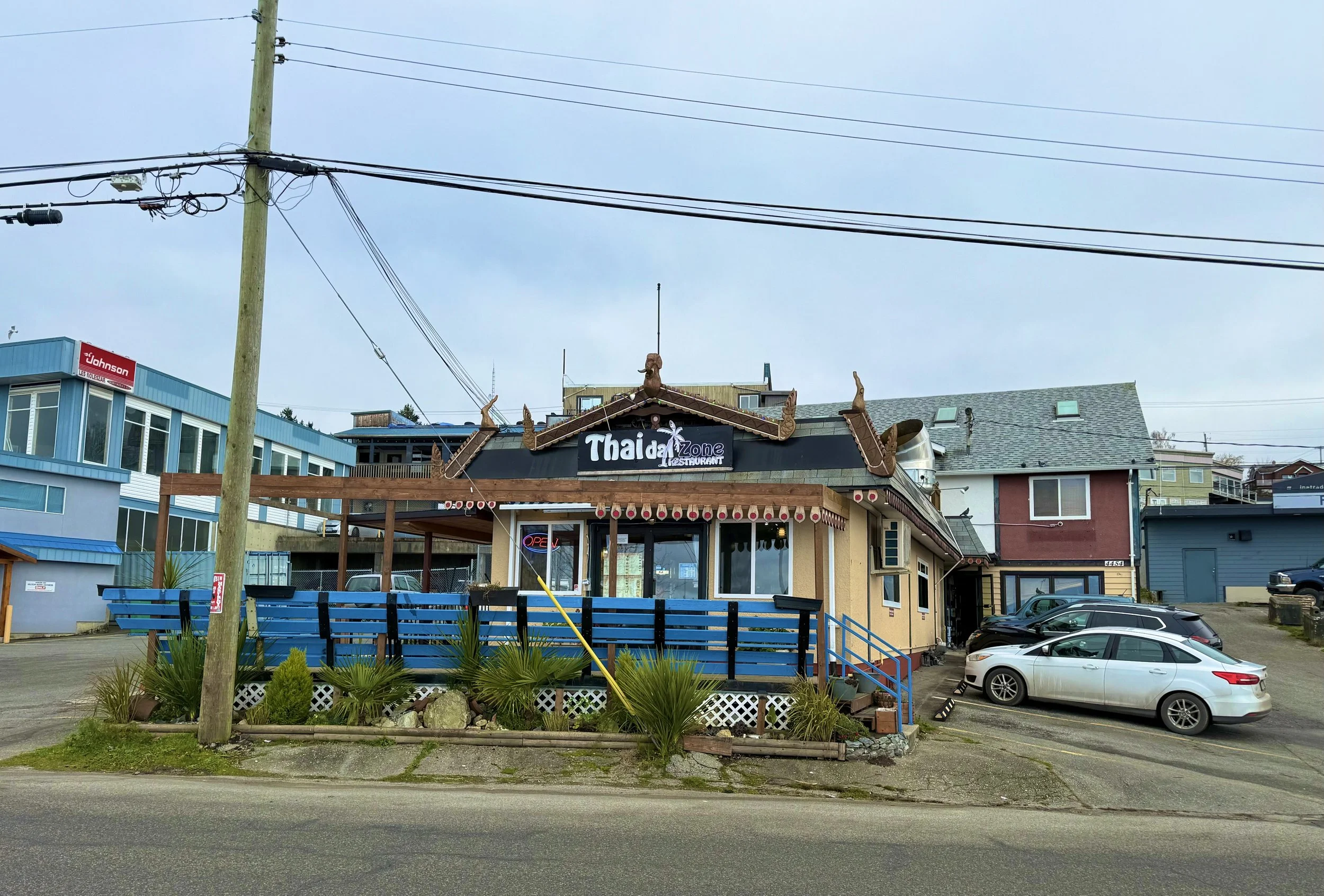 A Thai restaurant called 'Thai Zone' with traditional Thai architectural elements, including a tiered roof with decorative chofas, and a parking lot with several cars in front. The restaurant has a wooden deck with blue railings and potted plants out