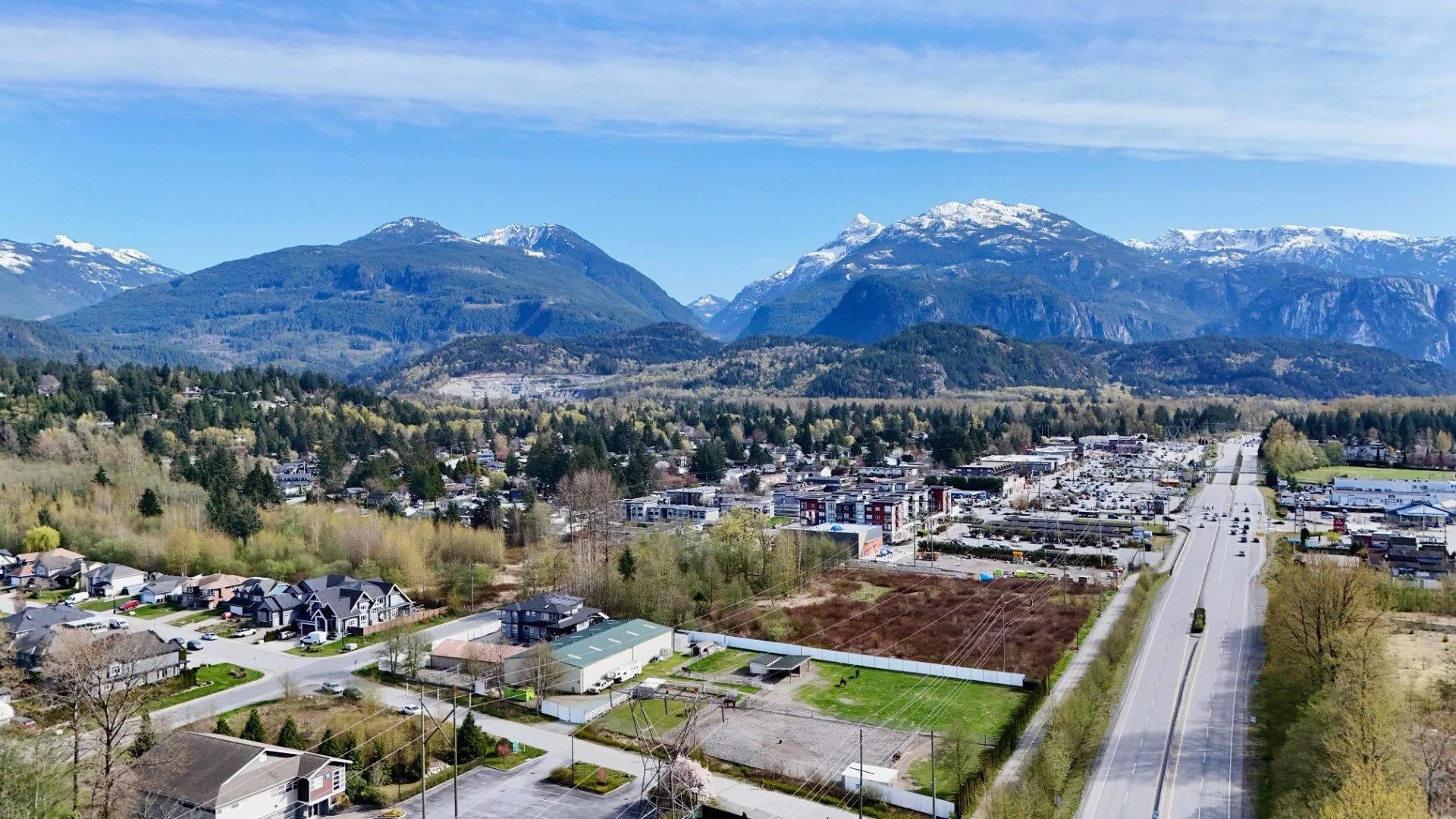 Aerial view of a small town with houses, commercial buildings, and roads, surrounded by green trees and mountains with snow on the peaks under a partly cloudy sky.