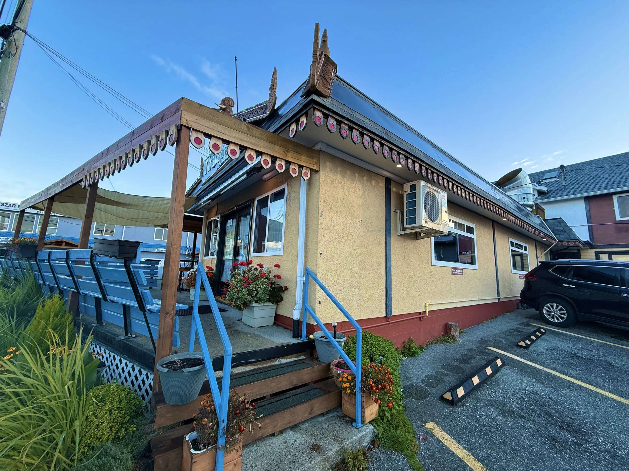 Exterior view of a yellow building with a small staircase and blue handrails, decorated with potted flowers and plants, and a black car parked in front, under a clear blue sky.