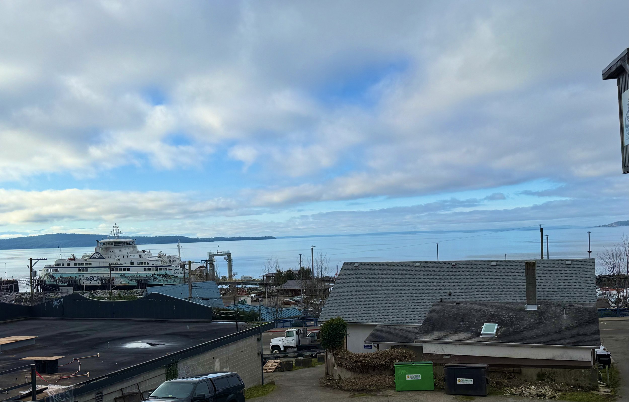 View of a harbor with a large white ferry boat docked, residential and commercial buildings in the foreground, and water extending to distant land with partly cloudy sky.