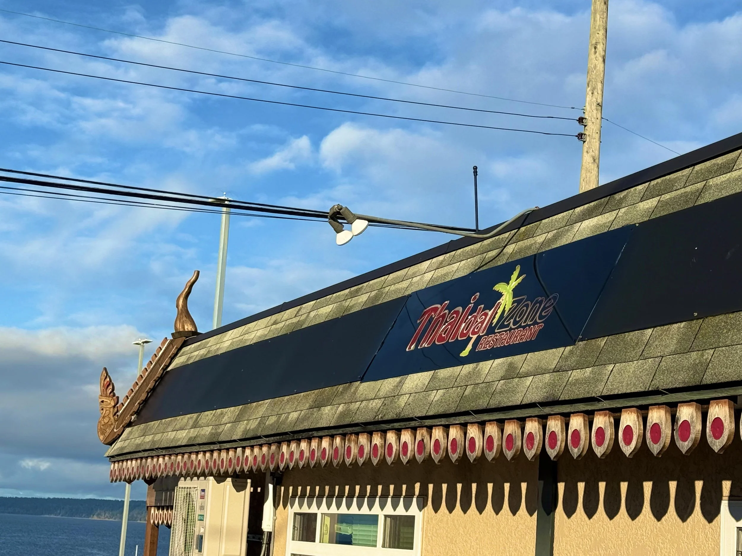 Sign for Thai Lad Zone restaurant on a building with a roof decorated with a dragon figure and a seagull perched on the roof, sky with clouds and power lines in the background.