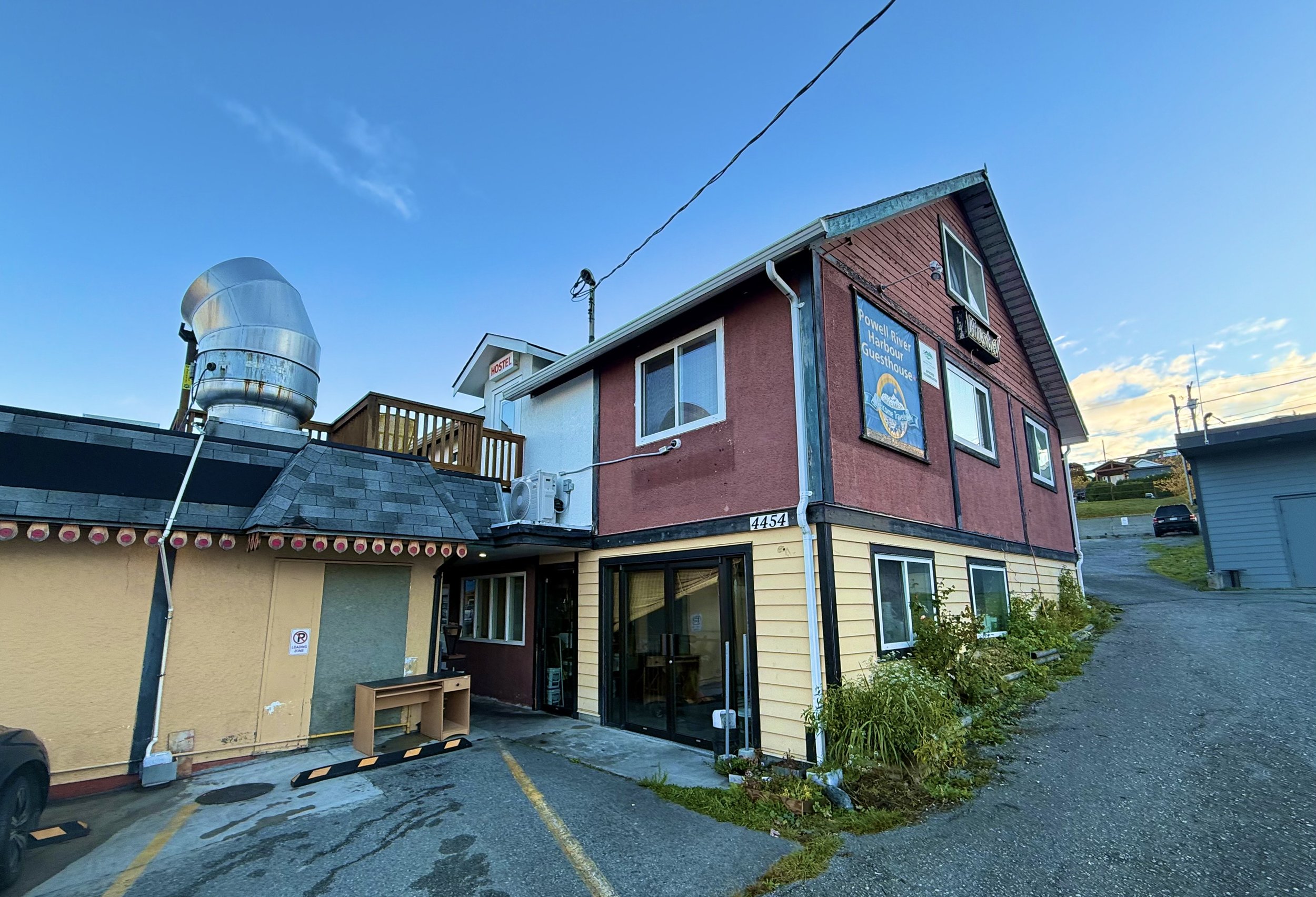 Multi-story building with signs for Powell River Harbour Guesthouse and Hostel, surrounded by parking lot, plants, and a gravel road, under a blue sky.