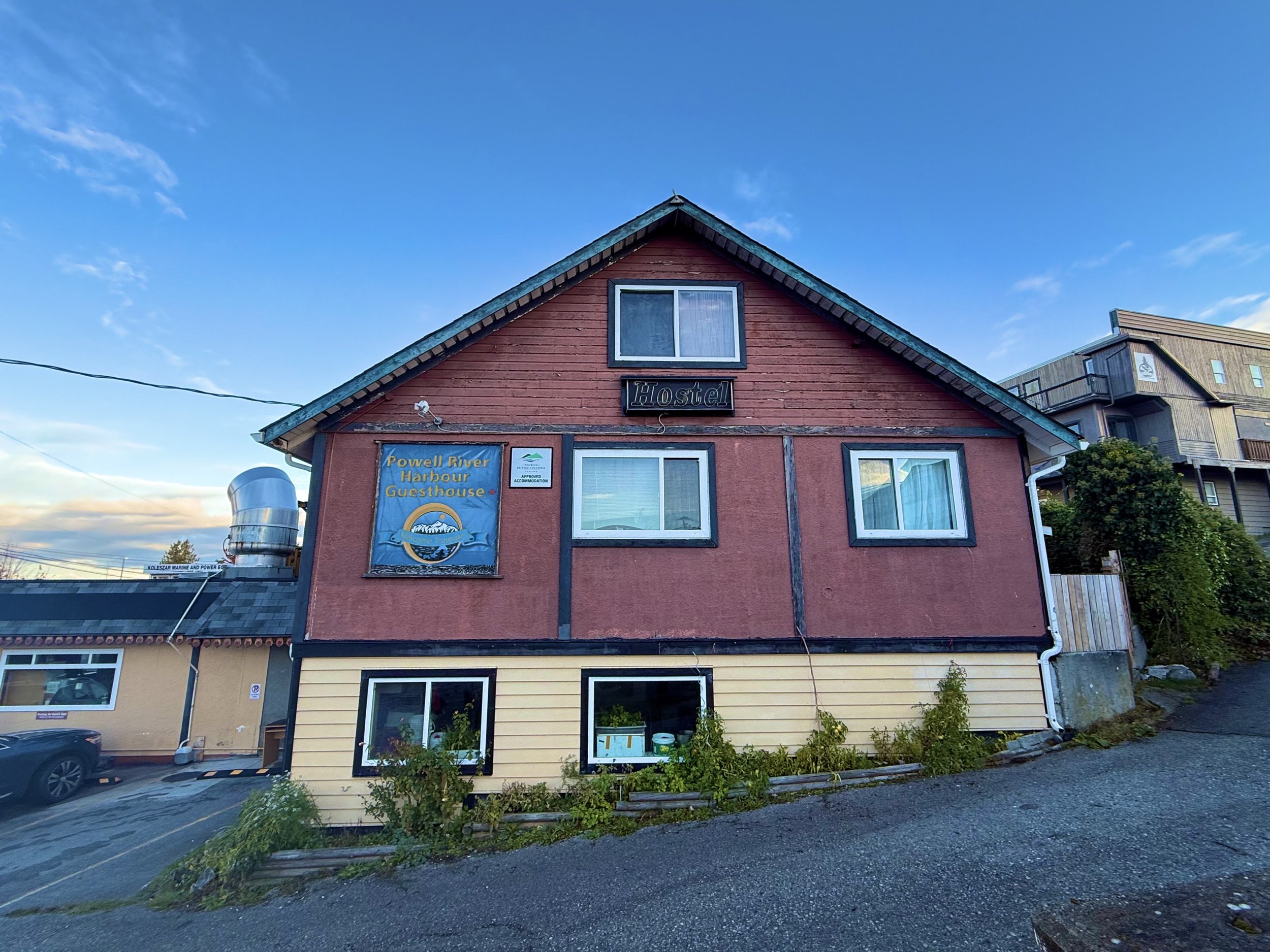 Two-story building with a sign that reads "Powell River Harbour Guesthouse" under a blue sky. The building is painted pink with black trim. The lower level has yellow siding and the upper level has wood paneling. There are three windows visible, two 