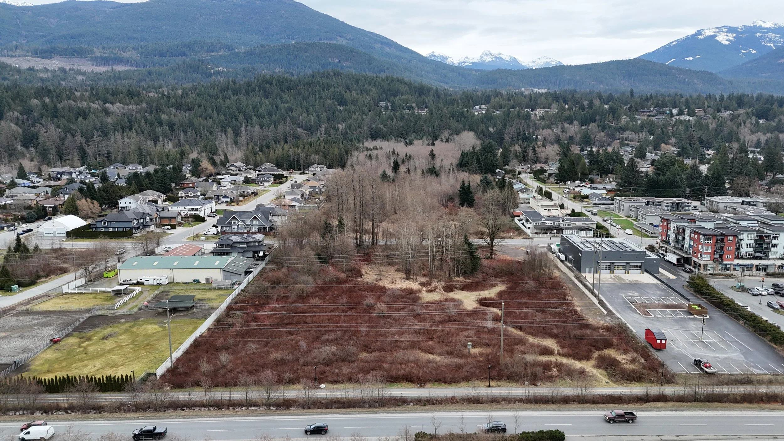 Aerial view of a small town surrounded by mountains with snowy peaks, featuring residential houses, a commercial building with parking lot, open land with trees, and a highway with cars at the bottom.