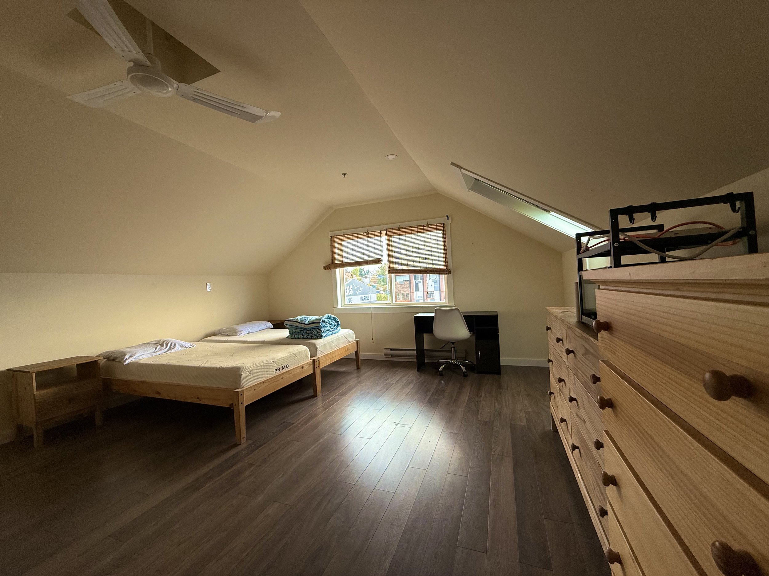 A bedroom with two single beds, a small wooden side table, a desk with a white chair, a window with bamboo blinds, a ceiling fan, and a wooden dresser, with wooden flooring and sloped ceiling.