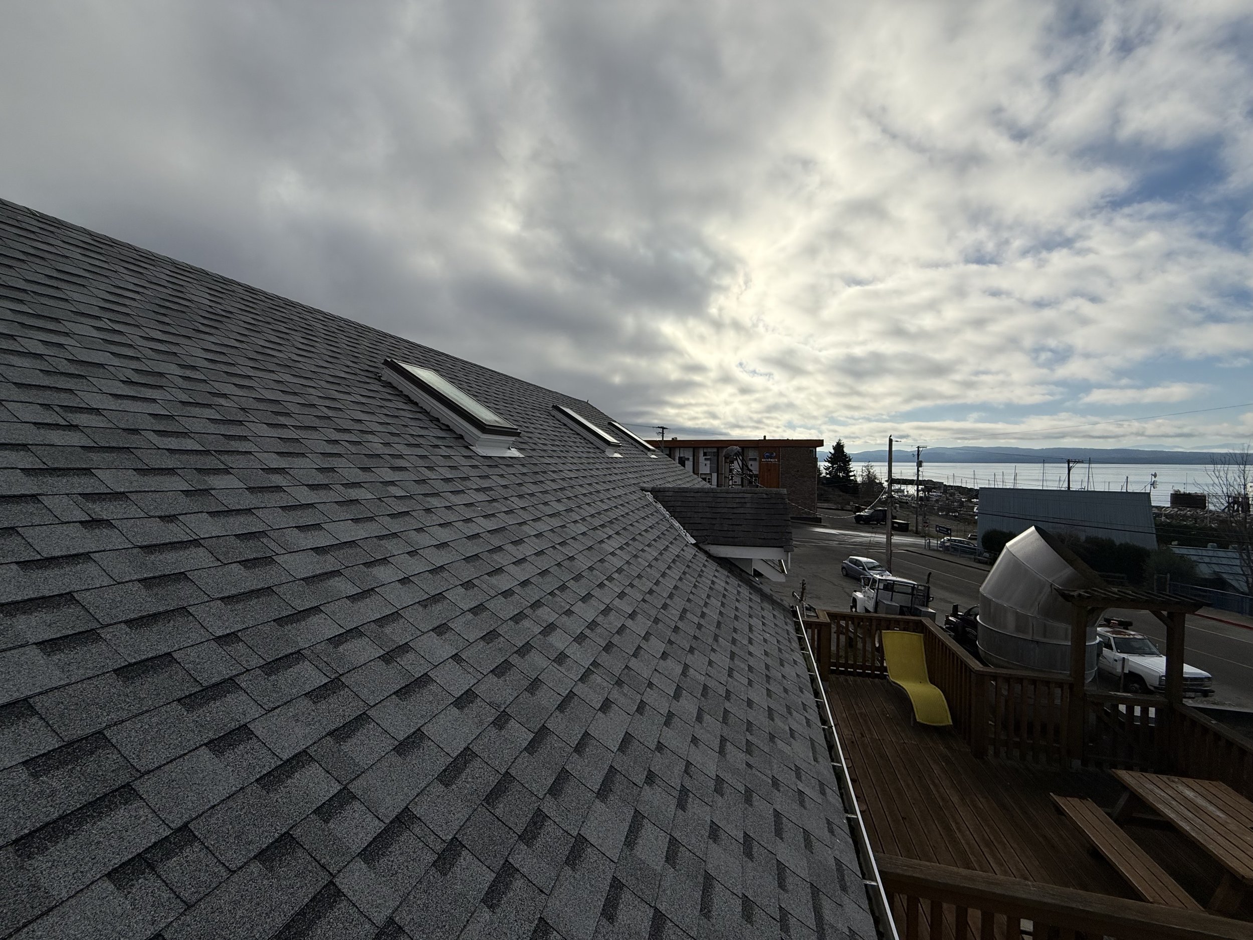 View of a rooftop with gray shingles and three skylights, overlooking a street with parked cars, buildings, and water in the distance under a partly cloudy sky.