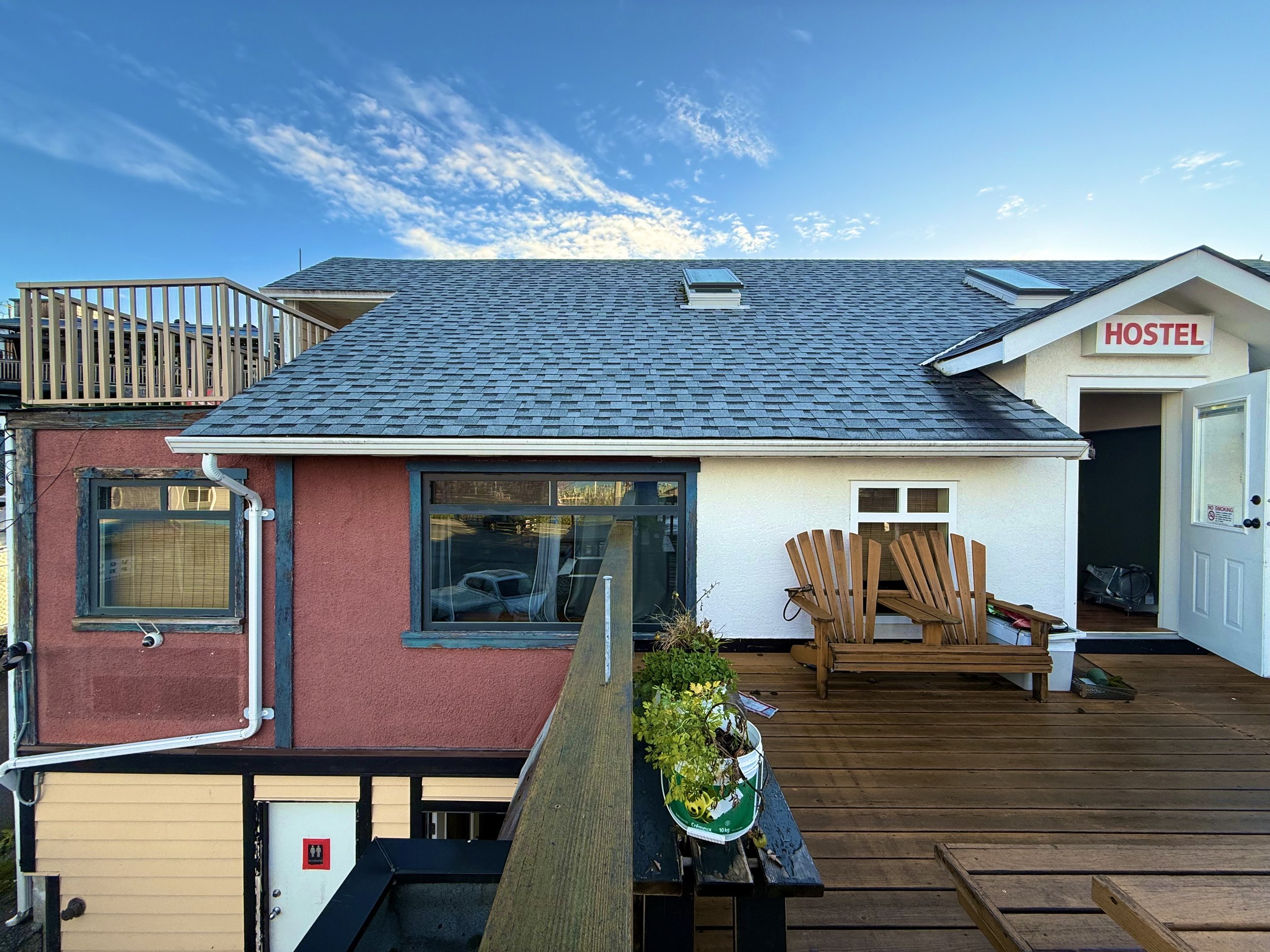 A rooftop deck with wooden flooring, a wooden Adirondack chair, a small window, and an open door leading to a hostel. The background shows a partly cloudy sky.