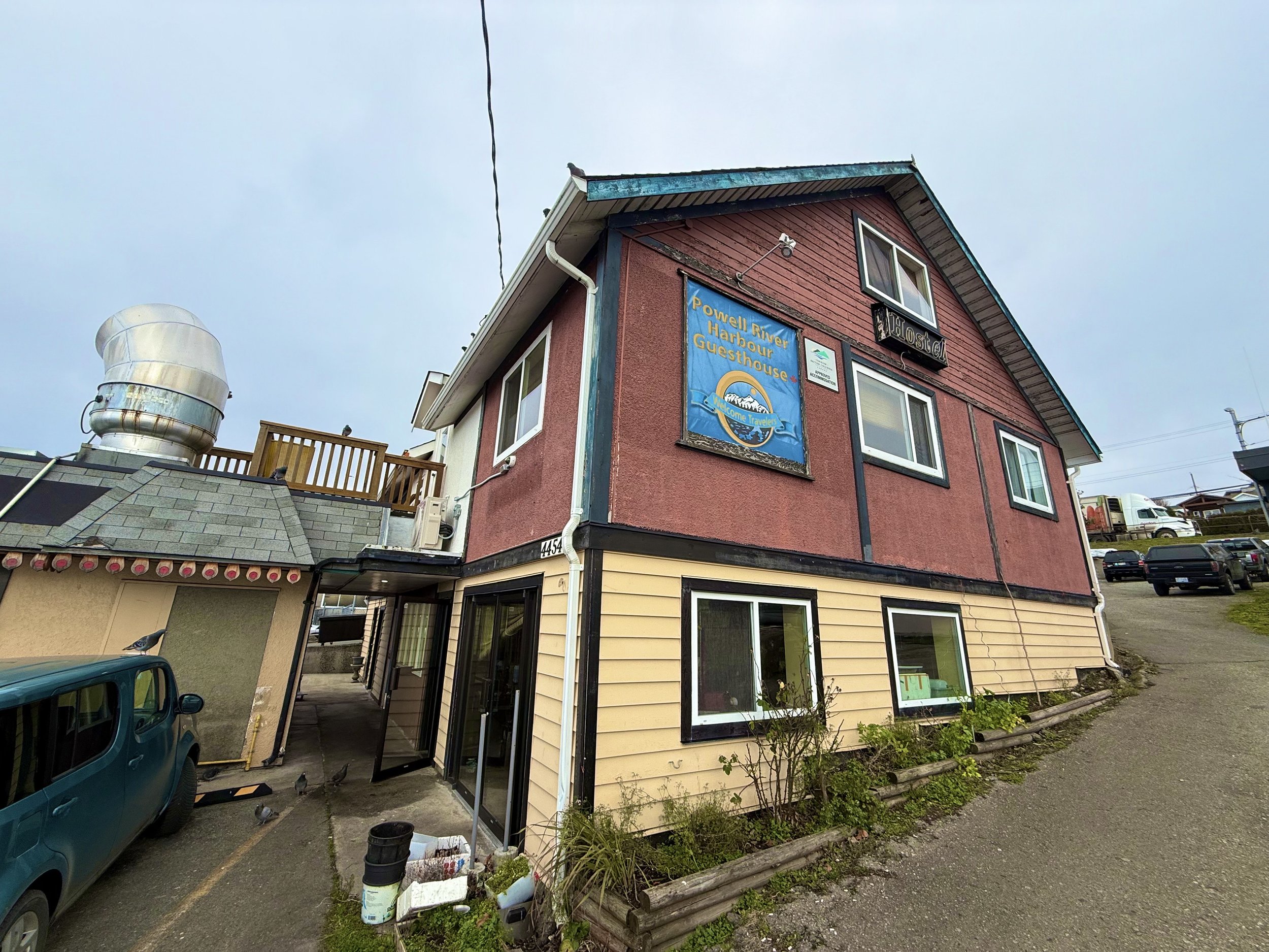 A three-story building with a garage and parking lot, blue sky, and a sign that reads 'Powell River Harbour Guesthouse.'