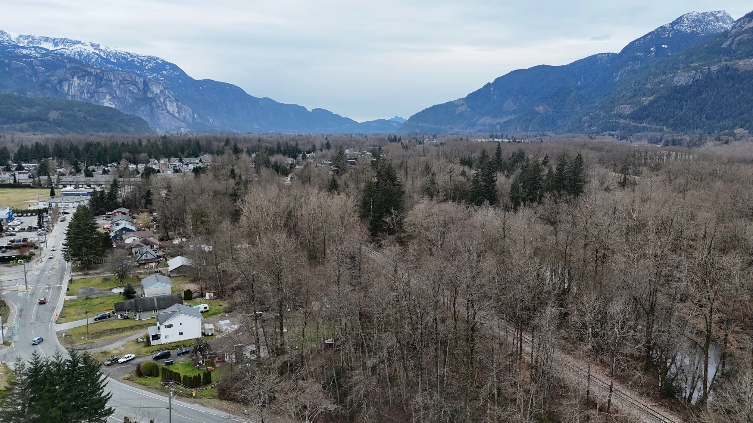 A scenic view of a small town with houses and cars, surrounded by leafless trees, with mountains in the background under a cloudy sky.