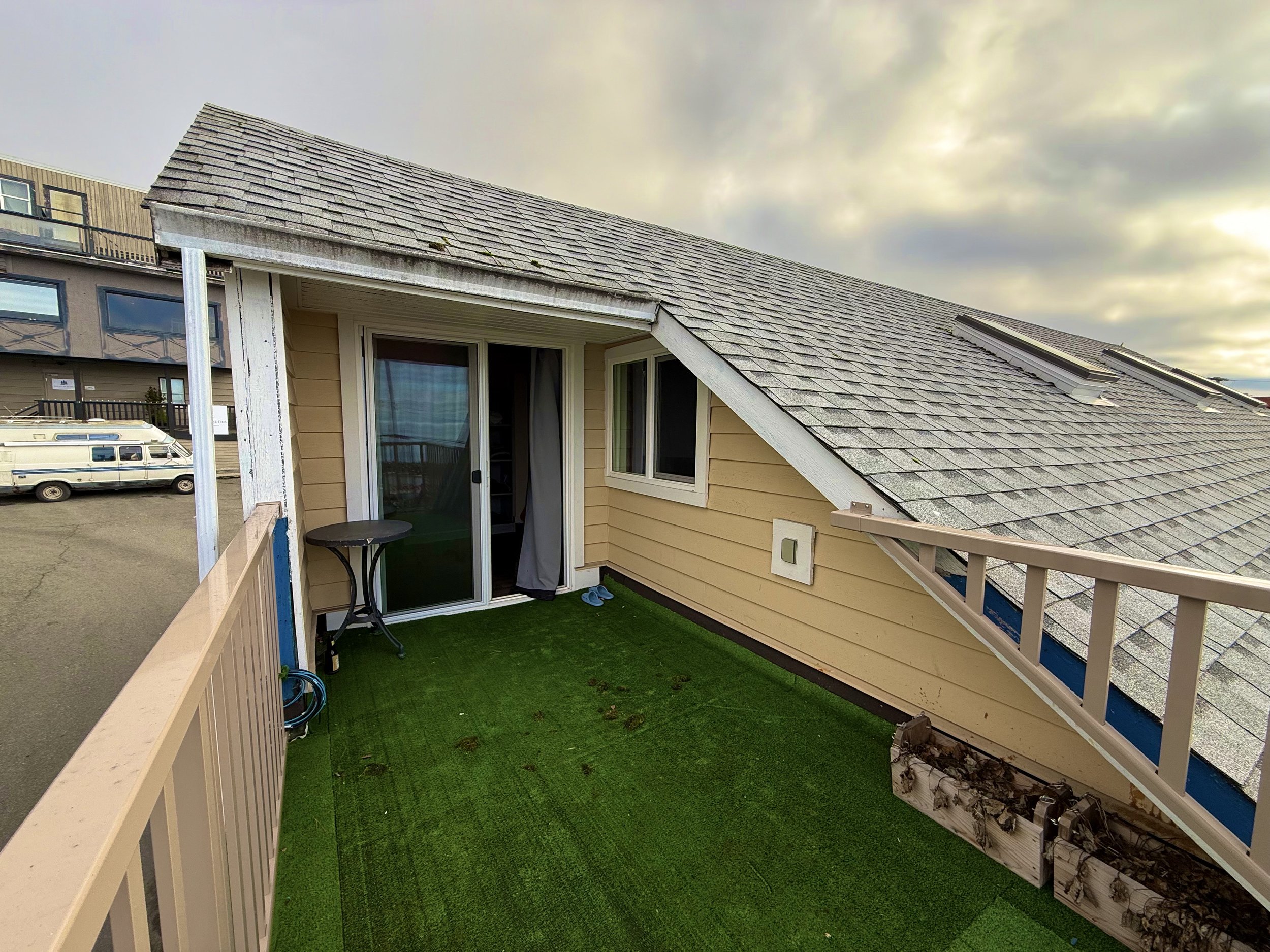 Small balcony with artificial green grass, a sliding glass door, and a window; beige siding; weathered roof shingles; cloudy sky; parked RV in the distance.