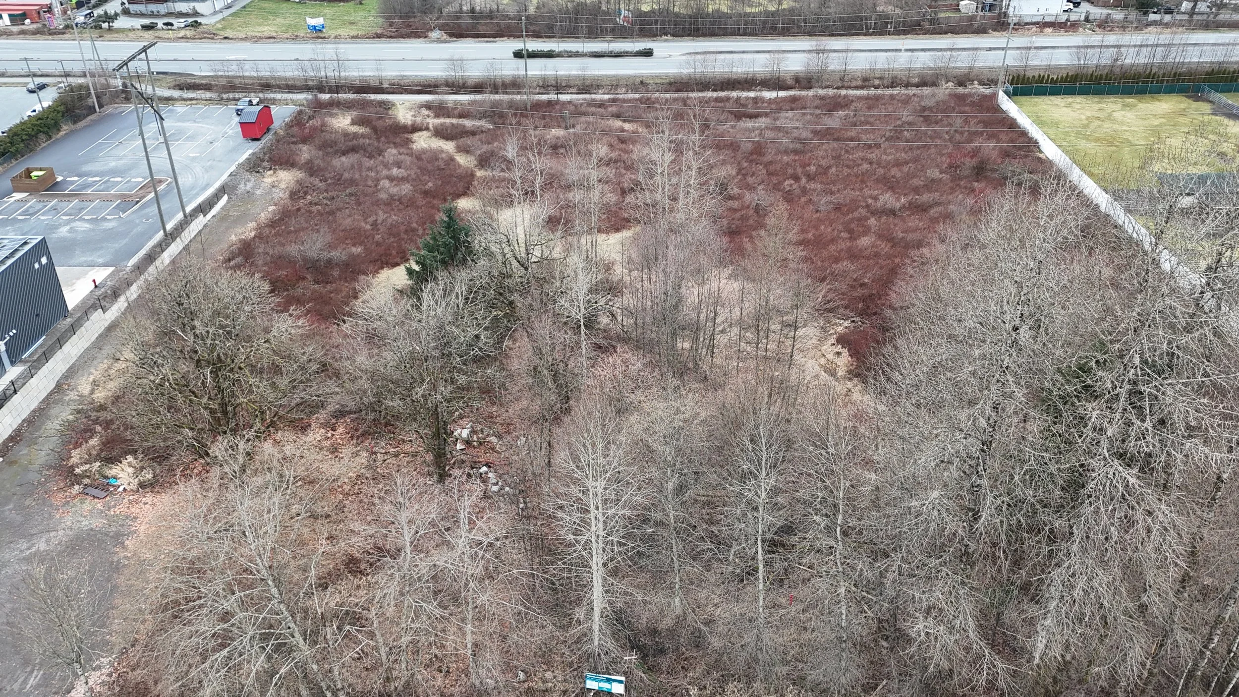 An aerial view of a vacant lot with leafless trees, some brown shrubbery, and patches of grass, bordered by a fence on the right side and a sidewalk on the bottom. A parking lot and road are visible in the background.