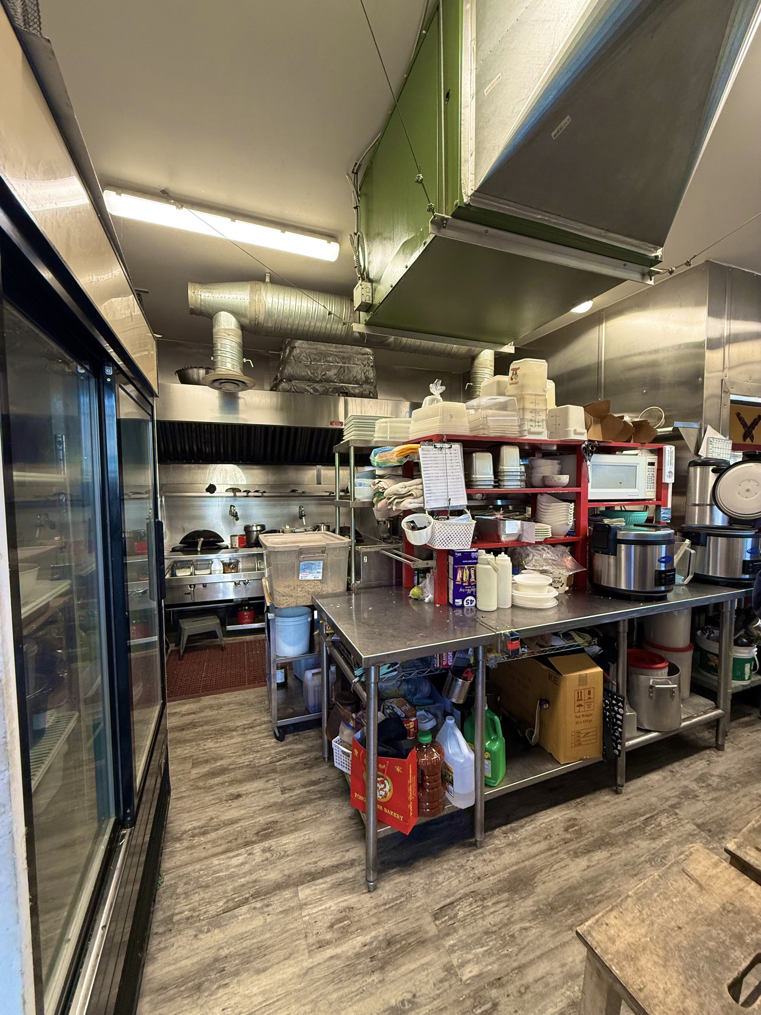 Commercial kitchen with stainless steel appliances, shelves, and storage containers, wood laminate flooring, visible ventilation duct and kitchen equipment.