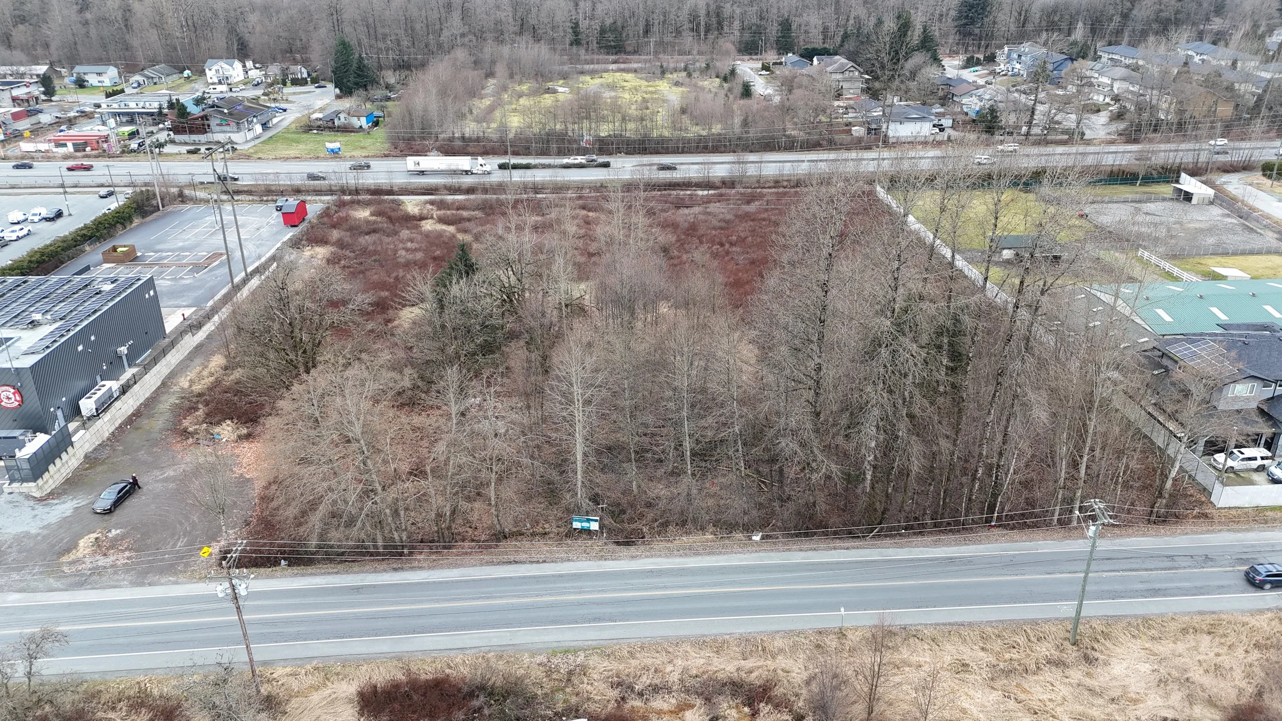 An aerial view of a wooded area beside a road, with residential houses and a parking lot in the background. The wooded area contains mostly leafless trees, indicating winter or early spring.