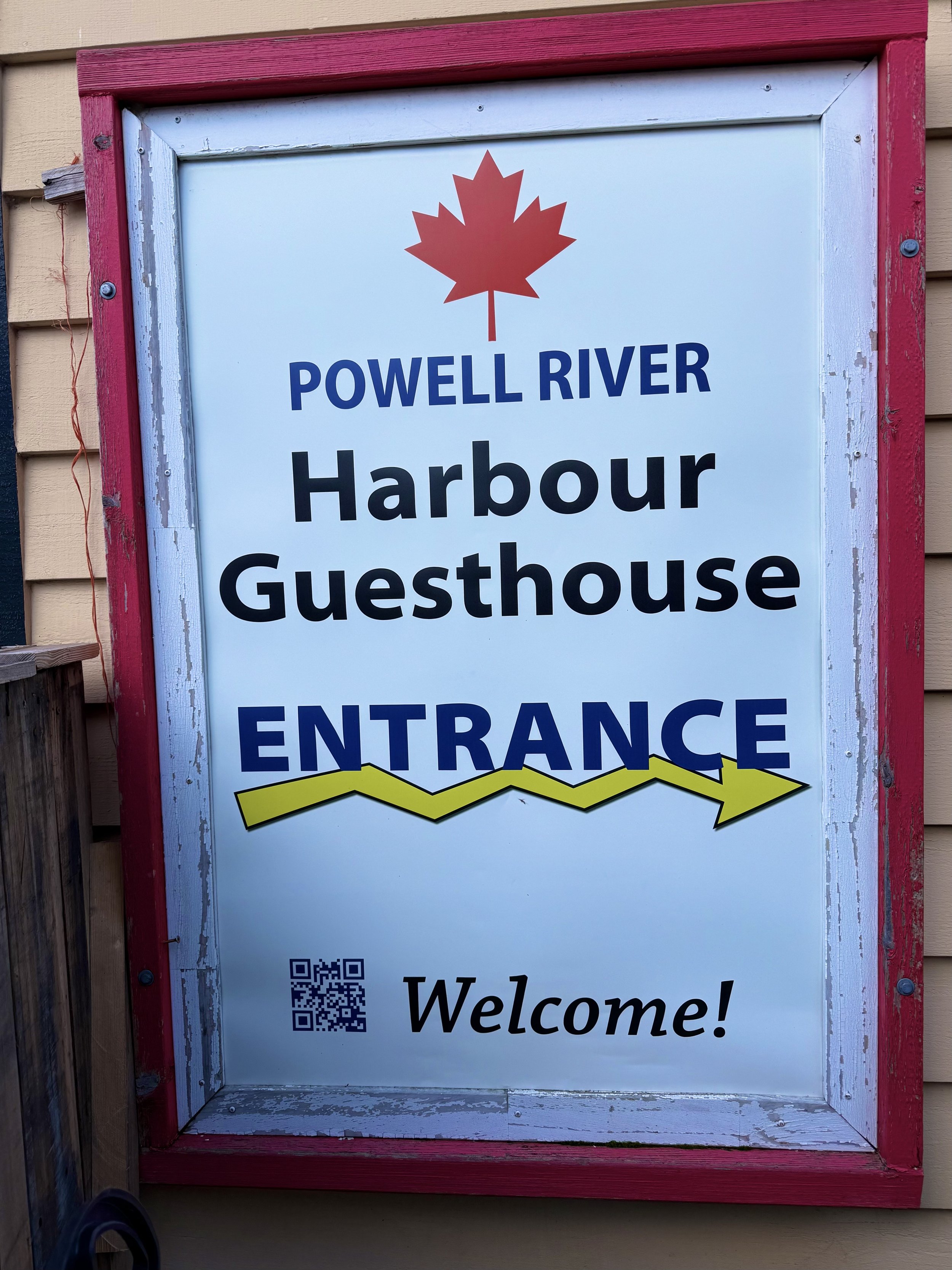 A sign for Powell River Harbour Guesthouse with a red maple leaf at the top, blue and black text, a yellow arrow, a QR code, and the word "Welcome!" at the bottom, mounted on a building with beige siding and a red frame.