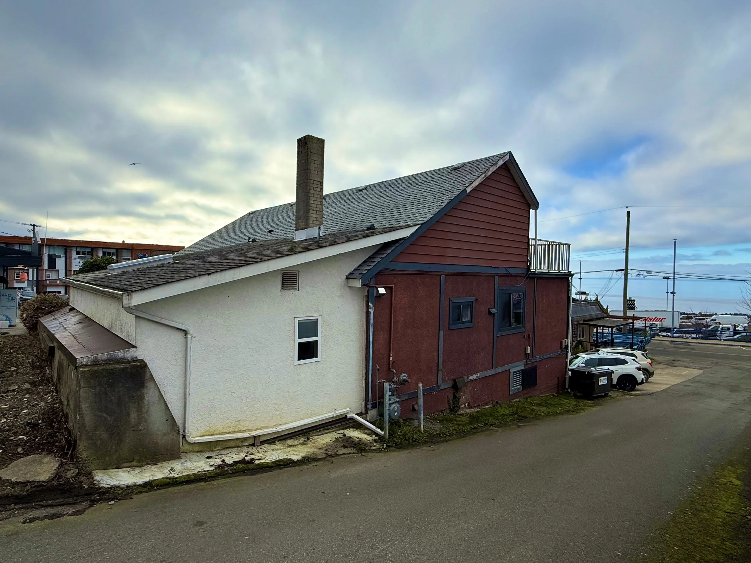 A two-story house with a gray shingle roof, a white lower section, and a reddish-brown upper section, situated next to a parking lot with several cars. The sky is partly cloudy with some sunlight breaking through.