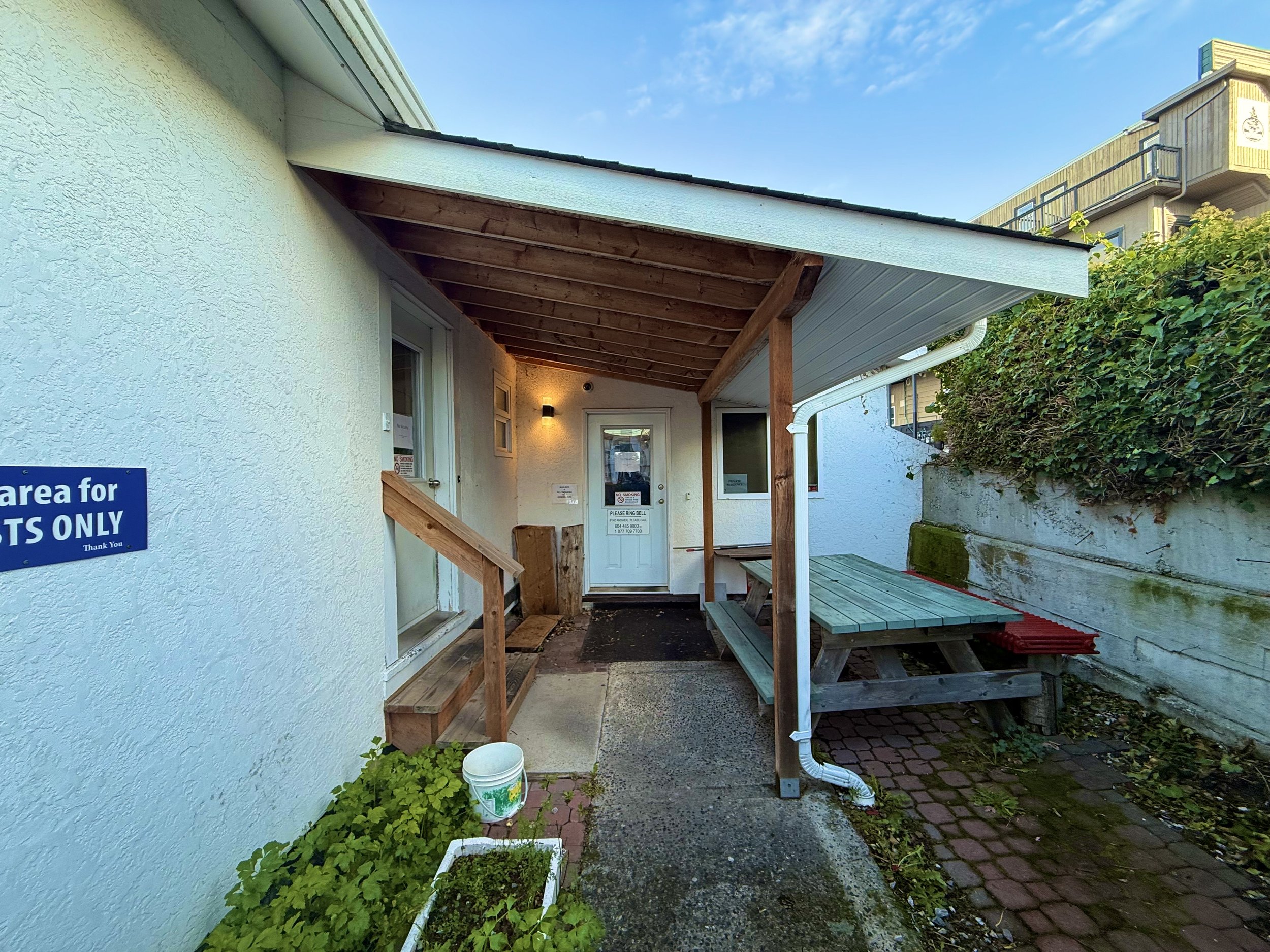 Small outdoor area with a sidewalk leading to a white door on a white stucco building. There is a wooden bench and picnic table under a wooden awning. Green plants in a white planter and a bucket are on the side, with a sign indicating a restricted a