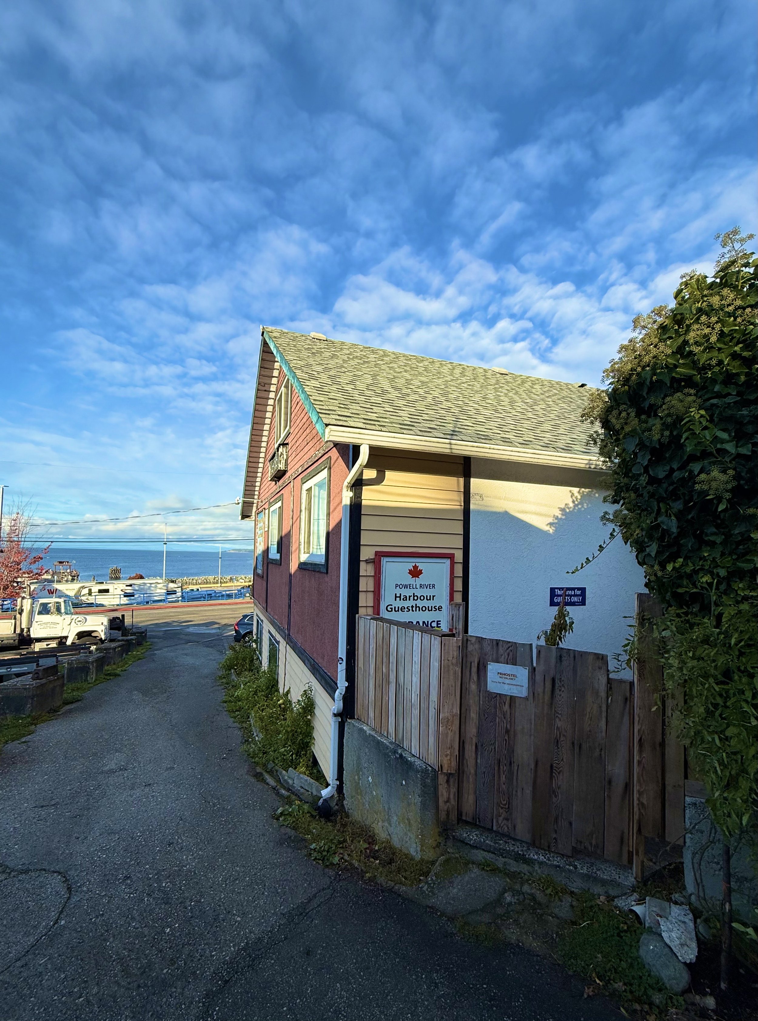 A seaside building with a pink and white exterior, sign indicating Powell River Harbour Guesthouse, and a gravel pathway leading towards the harbor with boats and water in the background. The sky is blue with some clouds.