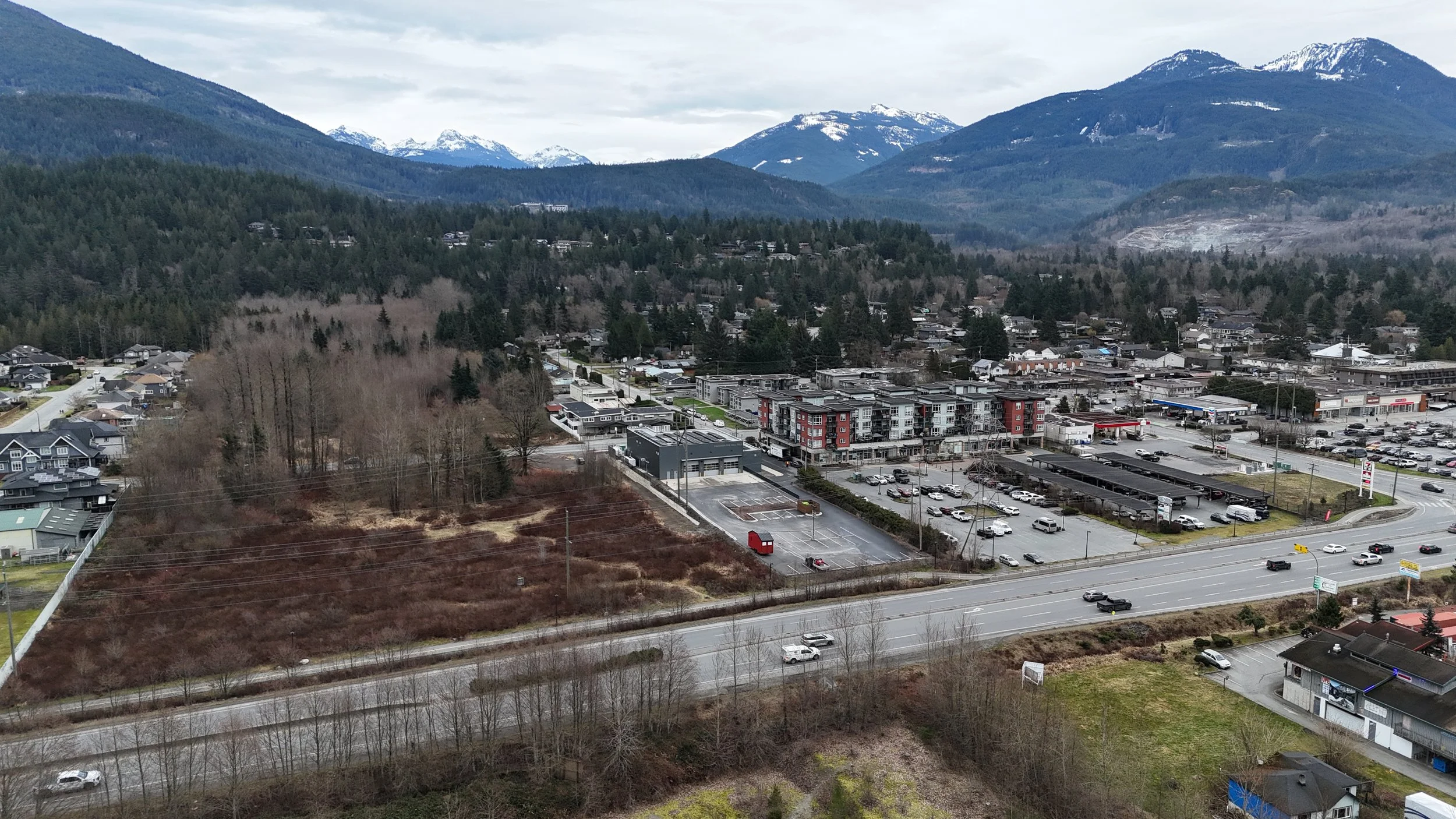 Aerial view of a town with a mix of residential and commercial buildings, a parking lot, and a highway with cars, set against a backdrop of forested mountains with snow-capped peaks under a cloudy sky.