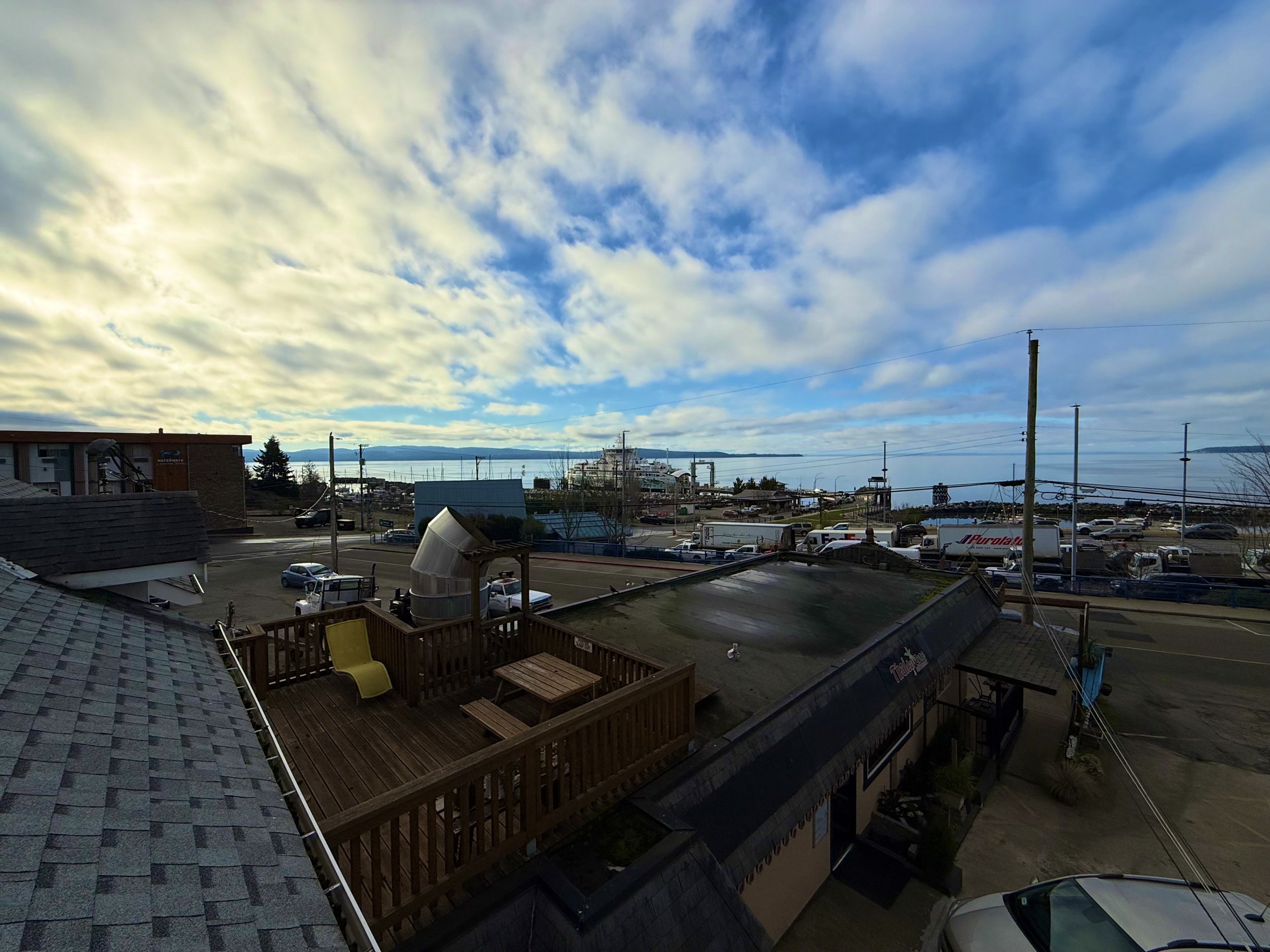 View of a parking lot, rooftops, and a waterfront with a large ship in the distance under a partly cloudy sky.